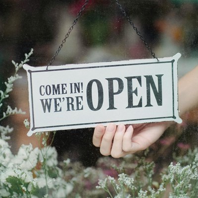 En hand som håller en skylt där står på engelska: Come in! we're open. Det finns blommor runt och under detta.