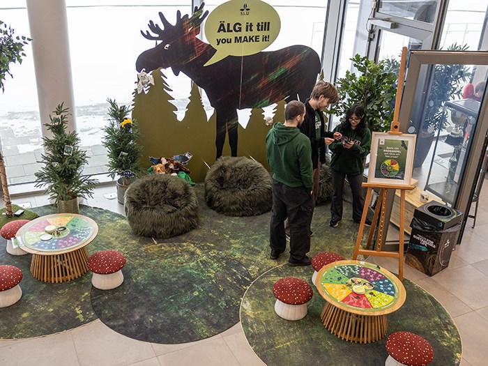 En monter är uppbyggd i en ljus lokal med stora fönster. Mitt i montern står en stor mörk silhuett av en älg med en gul pratbubbla som innehåller texten ”ÄLG it till you MAKE it!”. Framför silhuetten finns flera gröna, lurviga sittpuffar placerade på ett golv med skogsmönster. Till vänster och höger står gröna växter i krukor. I den högra delen av bilden står tre personer bredvid varandra och tittar på en mobiltelefon. I förgrunden syns två låga runda bord med svampformade pallar. Foto.