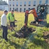 Two men watching a third man digging a hole with an excavator.