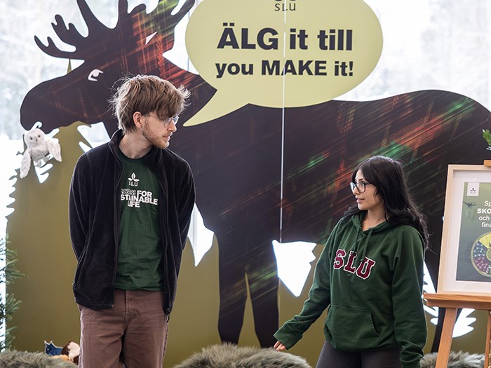 Två personer står framför en stor silhuett av en älg som är del av en monter. Ovanför älgfiguren finns en gul pratbubbla med texten ”ÄLG it till you MAKE it!”. Personerna står vända mot varandra och bär mörka ytterplagg och tröjor med SLU‑logotyper. Bakom dem syns älgsiluetten, dekor av gröna växter samt delar av en skylt på ett staffli. Foto.