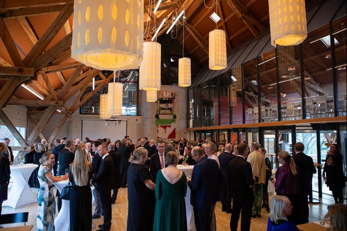 In a big hall with high ceilings, a large crowd is gathered mingling. Big white lamps hang from the ceiling.