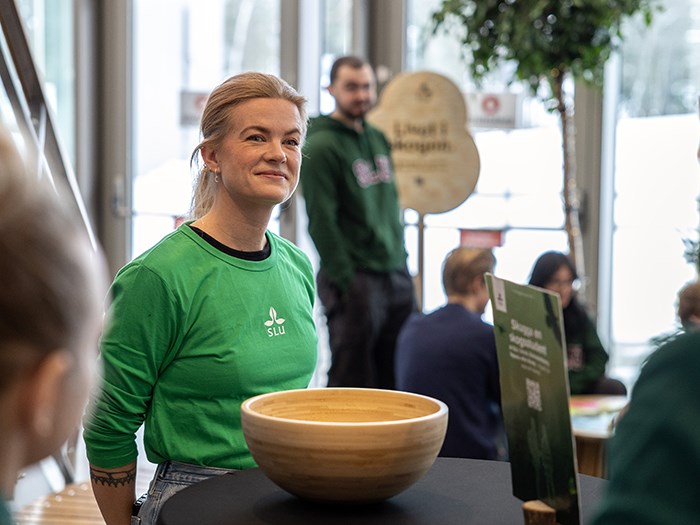 En person står bakom ett runt bord där en stor, ljus träskål är placerad. Personen bär en grön tröja med en SLU‑logotyp tryckt i vitt och har håret uppsatt. I bakgrunden syns flera andra personer i en ljus lokal med stora fönster, samt en träskylt med golvstativ och text samt ett trädliknande arrangemang. Några växter och delar av möbler kan också ses i rummet. Foto.