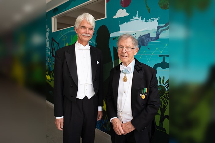 Two men in tail suits are standing side by side looking into the camera.