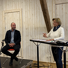 Man sitter på stol på scen. Kvinna står vid bord. Foto.