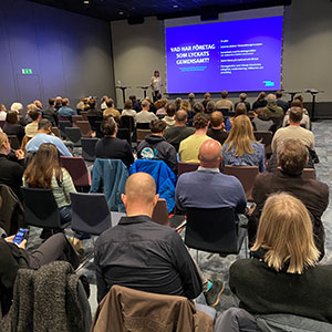 About 100 people at a conference listening to a presentation. Photo.