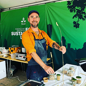 En person står vid en monter och håller upp två spannmålsstrån, ett kort och ett mycket långt. Foto. 