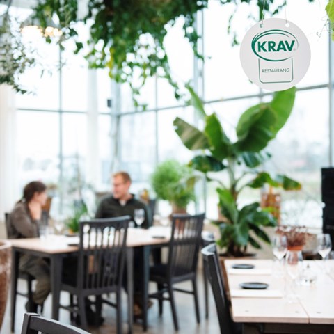 Interiörbild från en restaurang, väggarna består av fönster och det är många växter i lokalen. Från taket hänger KRAV:s logotyp.