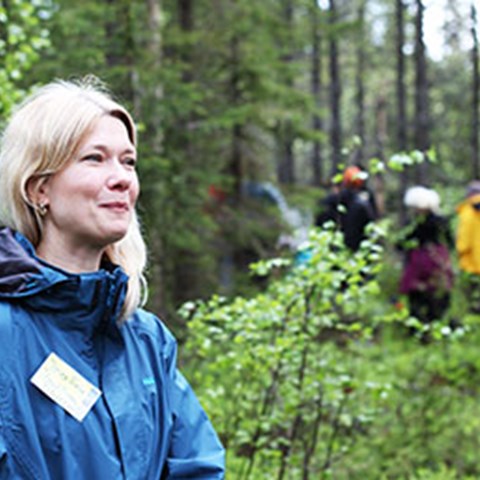Kvinna i närbild i skogsmiljö och i bakgrunden syns personer i oskärpa. Foto.