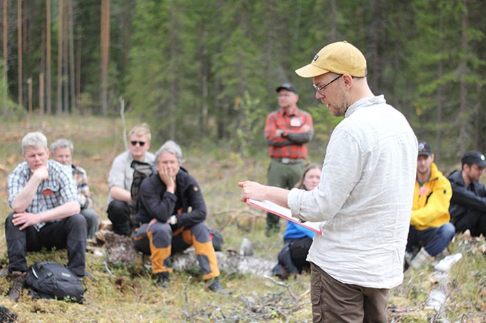 En man presenterar något och läser från ett papper framför en grupp människor som sitter i skogen på ett hygge. 