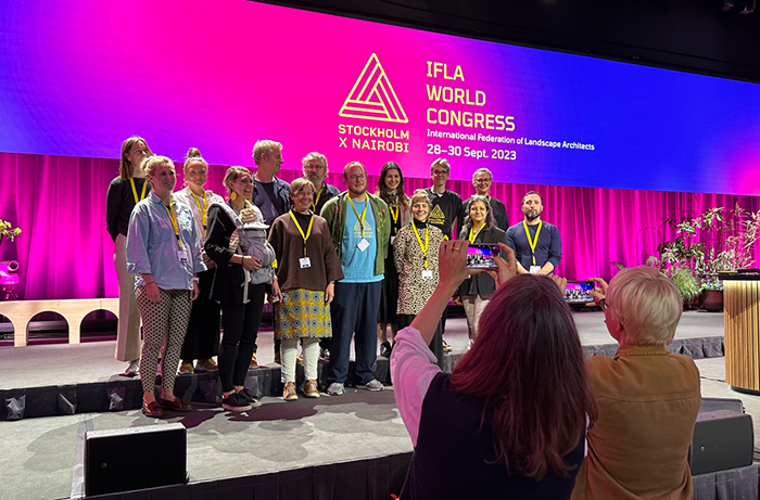 A group of people on a stage in front of a bright pink and blue background. People in the audience takes pictures. Photo.