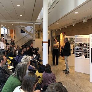 Audience listening to two presenters in front of poster exhibition.