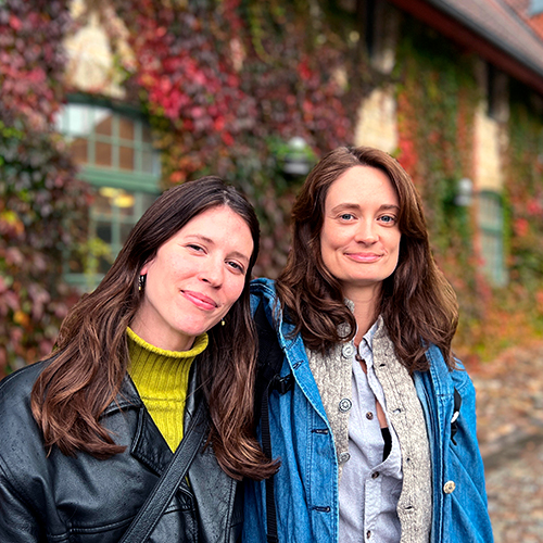 Två leende kvinnor står framför en tegelbyggnad täckt av grönska.