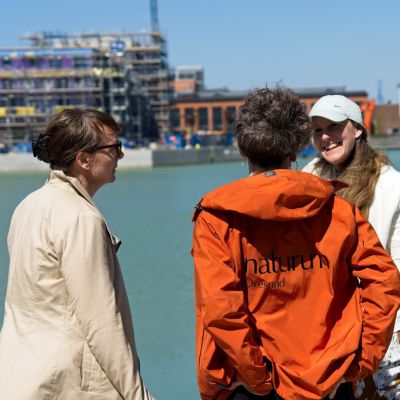 Tre kvinnor pratar med vatten och husbyggnation i bakgrunden.