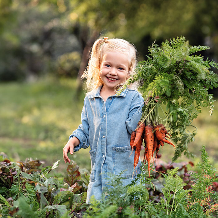 En leende flicka i blå klänning med ett knippe morötter i handen. Foto.