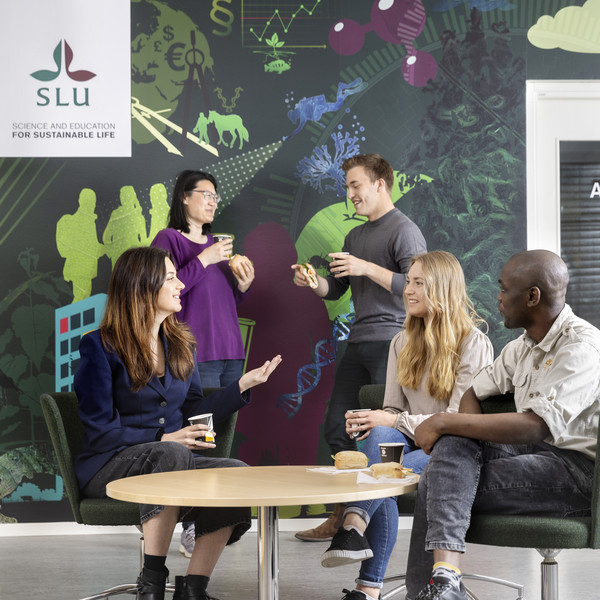 Studenter runt ett bord, i bakgrunden SLU-logotyp. Foto.