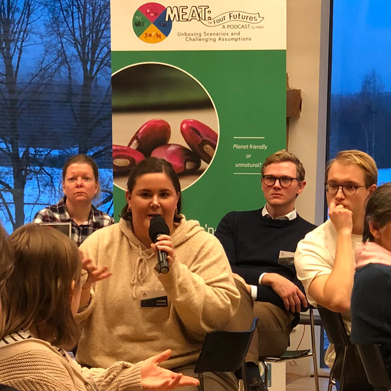 Workshop participants discussing. In the background a roll-up for the podcast Meat: the Four Futures.