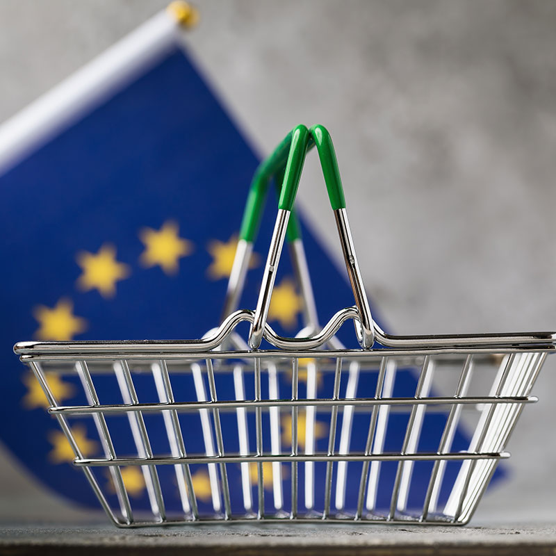 En kundkorg i miniatyr framför en EU-flagga. Foto.