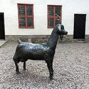 En getstaty i form av en get står på en grusad innergård. Foto. 