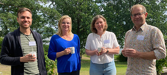 Two men and two women are standing outside, drinking coffee and chatting, with a glimpse of water and greenery behind them. 