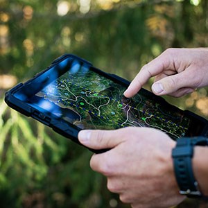 Ett par händer som håller i en Ipad, i bakgrunden skymtas en skog.Foto.
