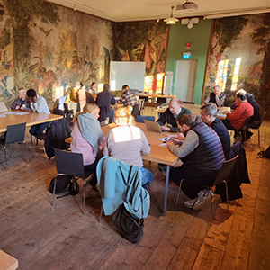 Personer sitter kring flera bord i ett rum med något som ser ut som antika målningar på väggarna.