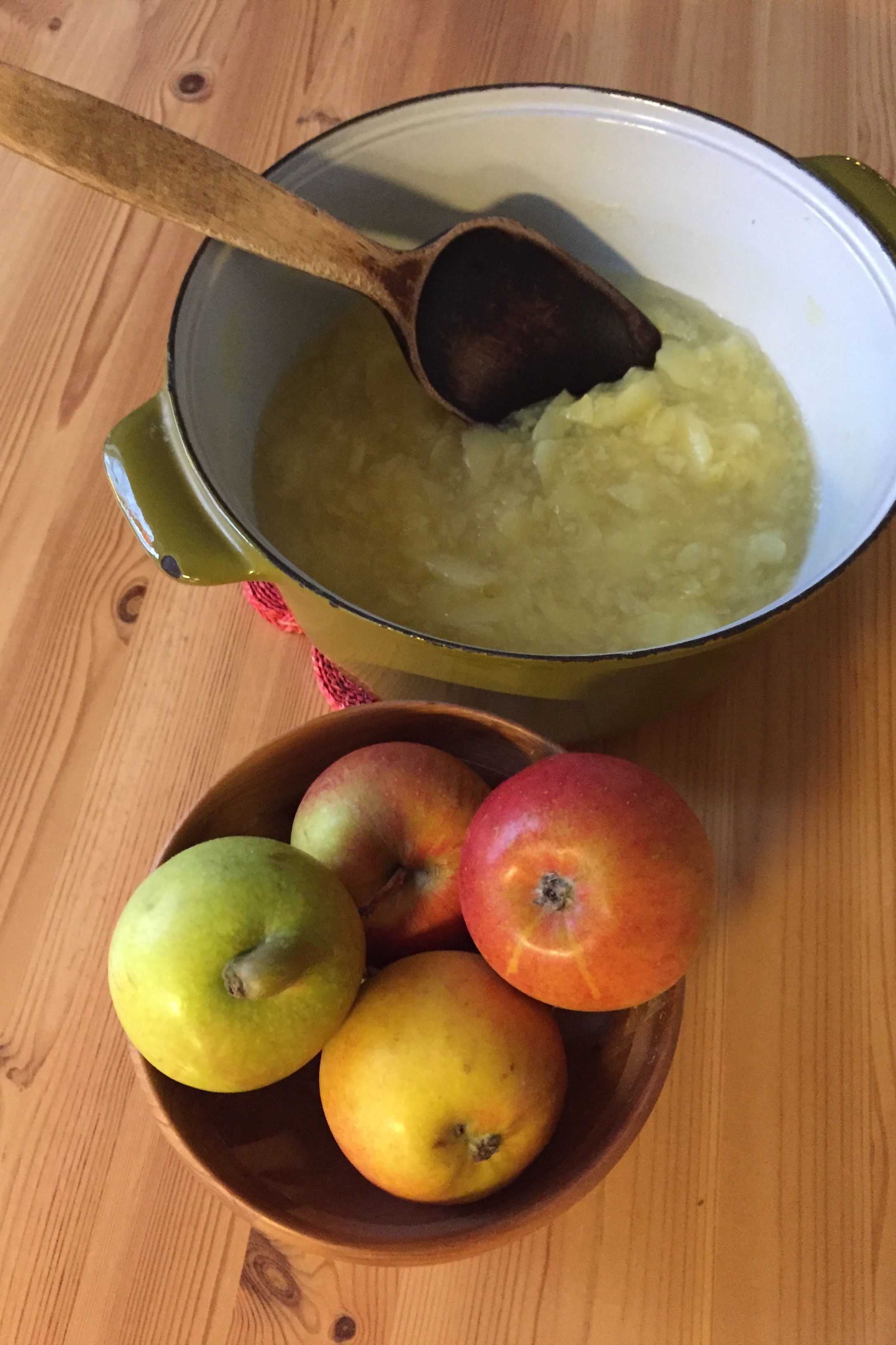 På ett bord står äppelmos i en grön gryta med en träslev i. Bredvid dem ligger fyra äpplen på ett fat. Färgfoto.