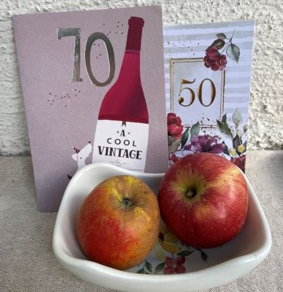 Äpplet Alfa 68 ligger till vänster i en skål. Till höger i samma skål ligger Aroma. Bakom Alfa 68-äpplet står ett födelsedagskort med siffran 70 och bakom Aroma syns ett födelsedagskort med siffran 50. 