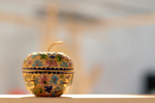 Fotot visar ett guldfärgat prydnadsäpple i metall, med dekor av blommor i blå och rosa emalj. 