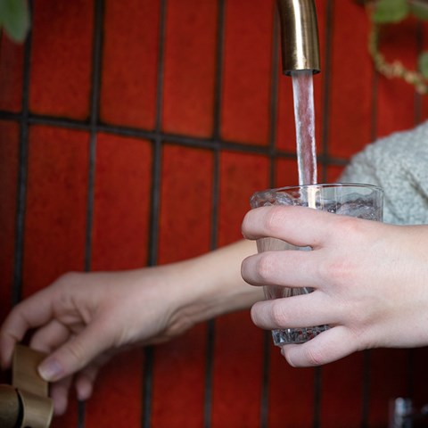 Two hands fill water in a glass from a tap. 