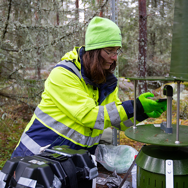 Person i tjock jacka arbetar vid en grön apparat i skogen.