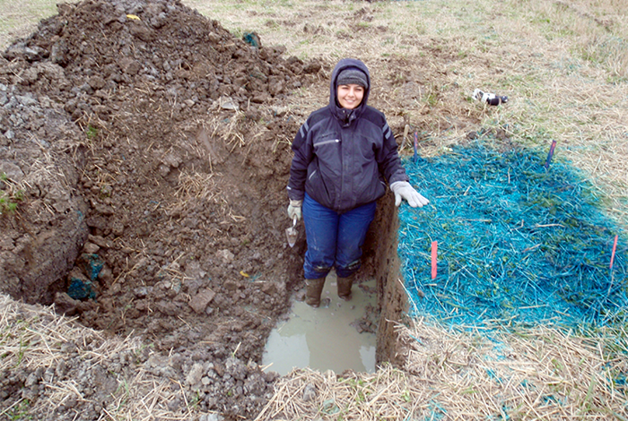 A woman on a field with a hole to the left. The ground to the right is colored blue. Photo.