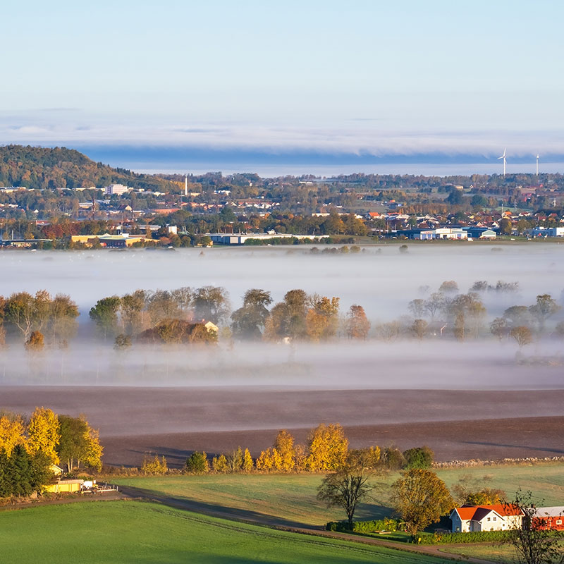 Dimstråk över landsbygd, i bakgrunden en stad. Foto.