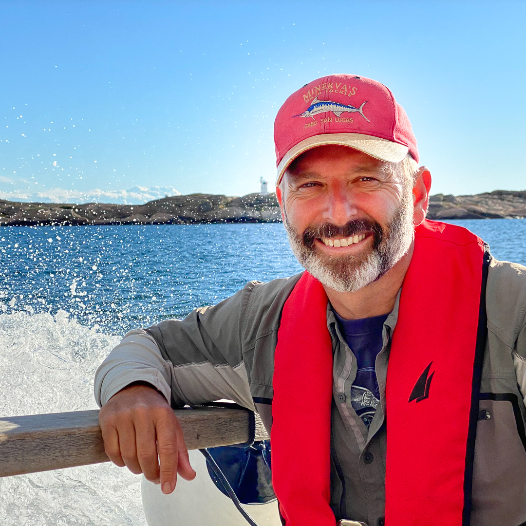 En man i flytväst och keps ombord på en båt. Foto.