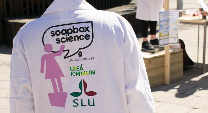 Rygg på vit labbrock med logotyper för Soapbox Science, SLU och Umeå universitet. Foto.