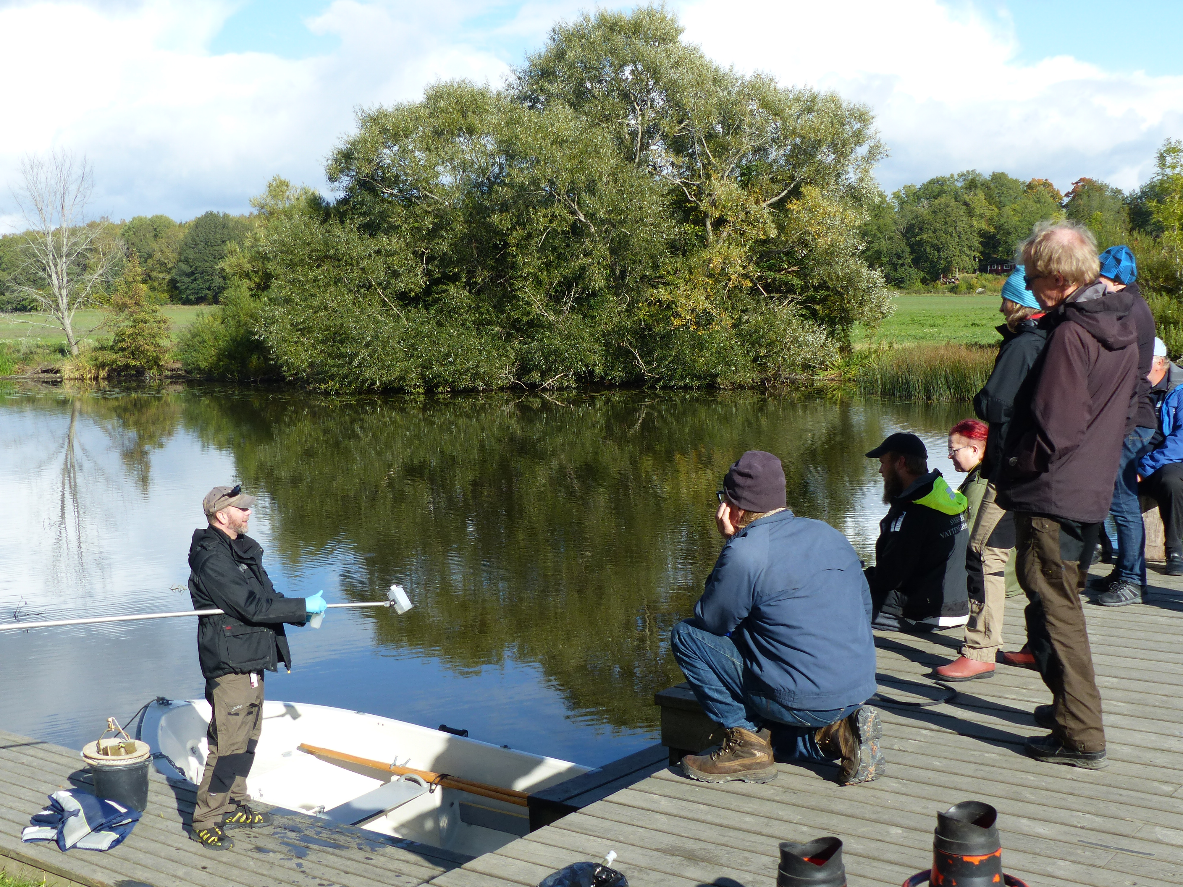Flera personer som står på en brygga och lyssnar på en man som visar hur man tar vattenprover. Foto.