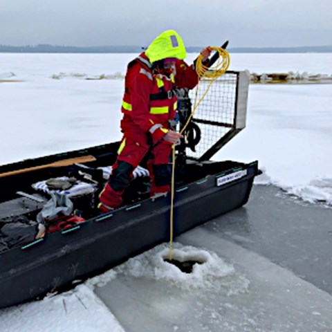 Man som står i hydrokopter och sänker ner utrustning i borrat hål i isen. Foto. 