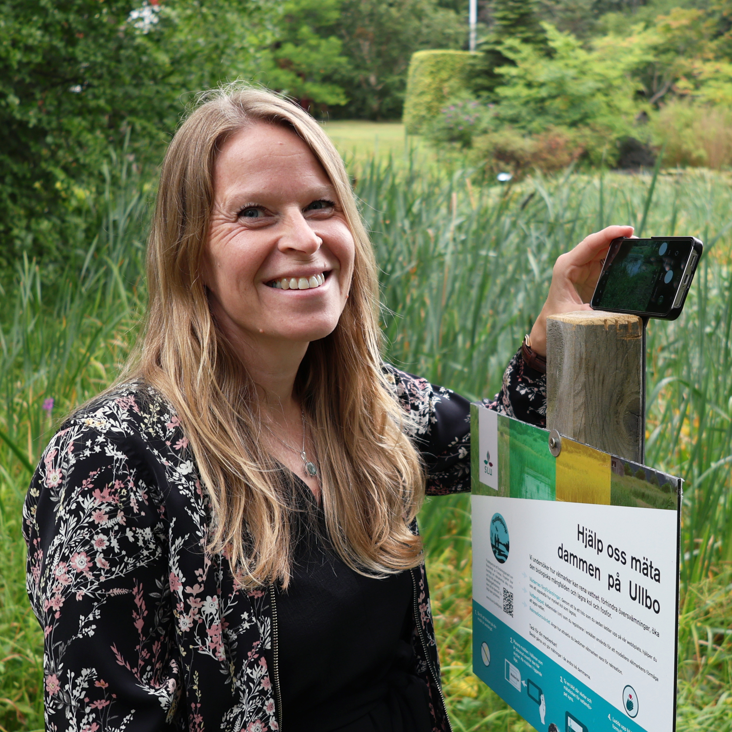 Kvinna håller mobiltelefon vid skylt framför en damm. Foto.