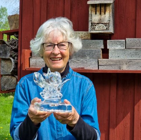 Kvinna med blå skjorta och grått hår håller i en glasstatyett. Foto