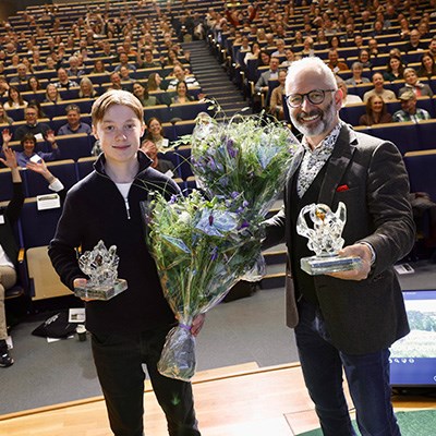 Man och pojke leendes med pris-statyetter och blombuketter framför stor publik. Foto