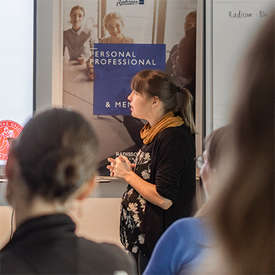 Kvinnlig talare står i profil bredvid projektorduk och syns inramad mellan åhörares huvuden. Foto