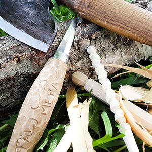 En yxa och en täljkniv lutade mot en trädstam, med små bitar trä, fotografi.