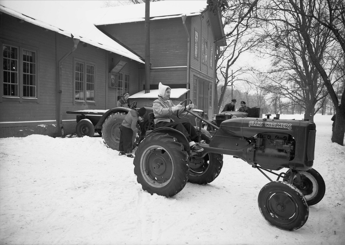 Traktor bredvid hus i snö, svartvitt fotografi. 
