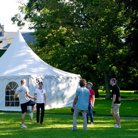 An image of five persons standing in a circle. One of them has a football. A tent in the background. Summer.