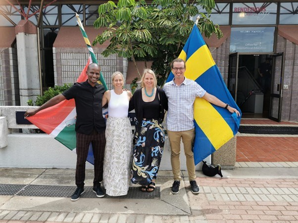 Four people standing with the Swedish flag and the South African flag behind them