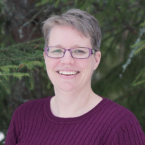 Leende kvinna står framför granar. Foto.