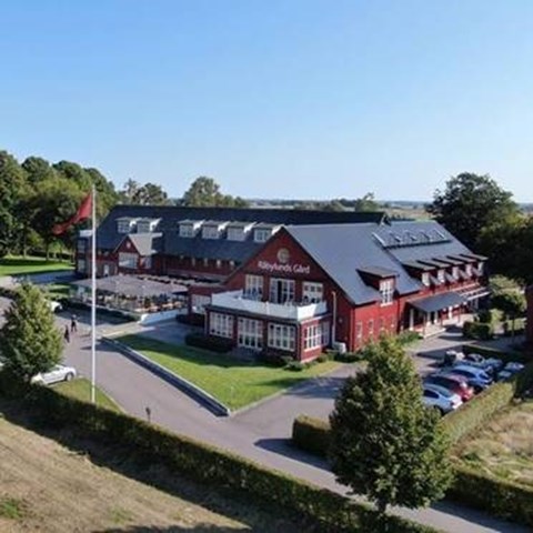 På bilden ser man Råbylunds gård, med röda byggnader, svarta plåttak, parkerade bilar, människor i rörelse och en uteplats i lummig grönska.