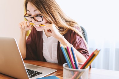 En person sitter vid ett träskrivbord och arbetar på en öppen bärbar dator. Personen bär glasögon, en vit topp och har långt hår och biter på en penna. På skrivbordet finns en behållare med flera färgpennor. Foto.