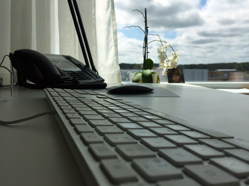Tangentbord, mus och telefon på ett vitt skrivbord. I bakgrunden ett fönster där man ser blå himmel med moln. Foto.