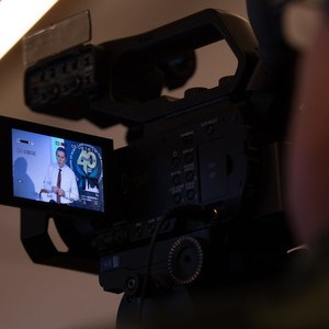 Picture of a video camera. In the viewfinder you see a man giving a lecture.