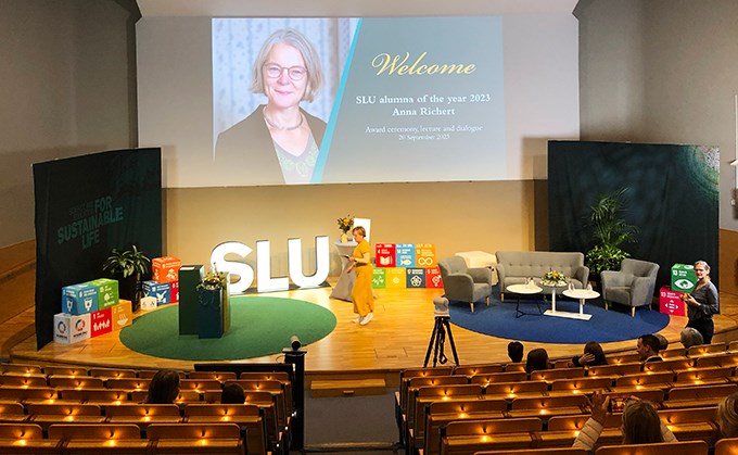 På en scen finns runda gröna och blå mattor, lysande SLU-bokstäver, sittmöbler och en projicerad bild av årets alumn.